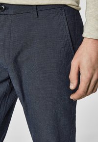 Dark blue trousers made from textured fabric, featuring a button closure, side pockets, and a tailored fit, displayed with a hand near the waist.
