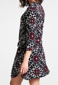Černo-bíl dresses s vzorem a bordo akcenty. Má dlouhé rukávy a volný střih, vyrobené z lehkého materiálu.