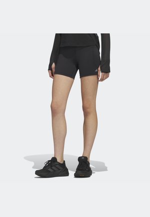 Personne portant un short de sport noir et des chaussures de course noires, se tenant sur un fond blanc avec une ombre en dessous.