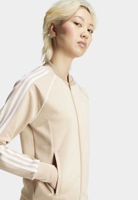 Chaqueta beige con cremallera, confeccionada en tela suave, con acentos en blanco de tres rayas en las mangas y bolsillos laterales.