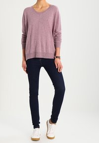 Jasnoróżowy, oversize'owy sweter z okrągłym dekoltem, zestawiony z ciemnymi, dopasowanymi dżinsami i białymi trampkami z beżowymi podeszwami, stojący na neutralnym tle.