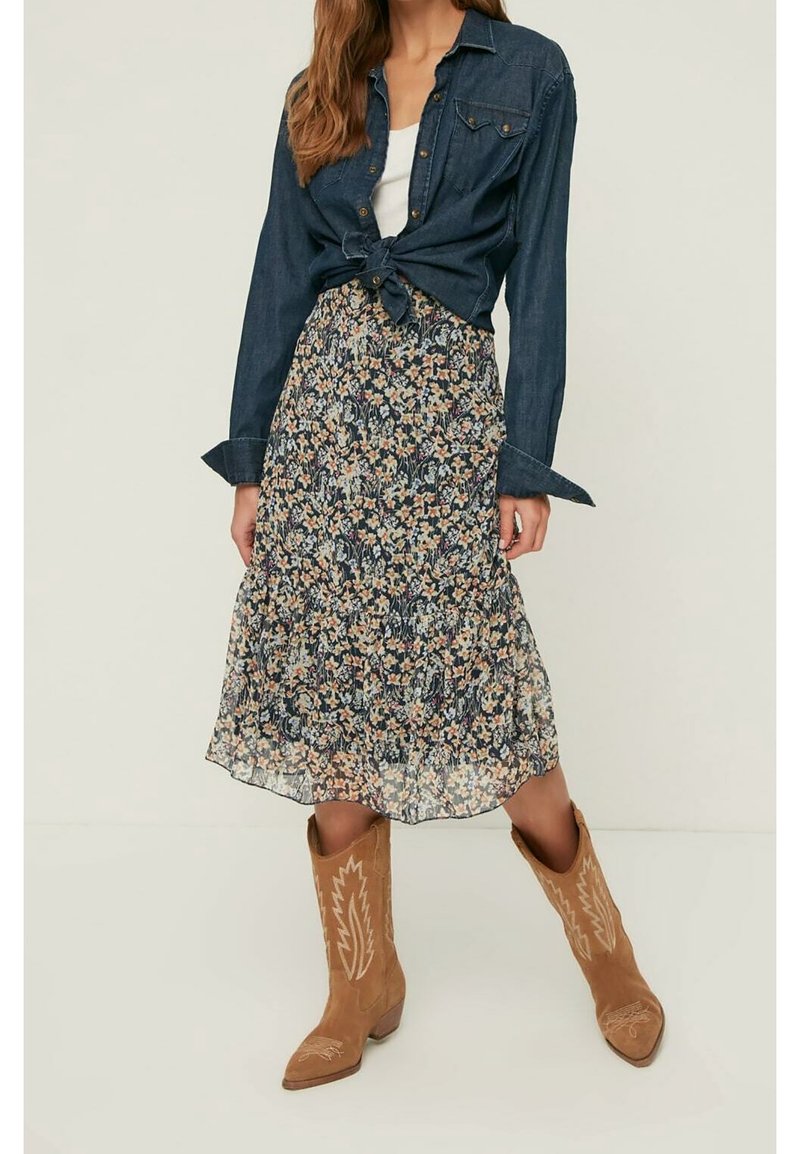 Frau trägt ein gebundenes Jeanshemd, weißes Oberteil, geblümten Midirock und hellbraune Wildleder-Cowboystiefel mit gestickten Mustern.