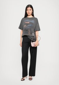 Tricou gri supradimensionat cu un grafic de benzină, asortat cu pantaloni larghi negri. Ține în mână o geantă mică roz. Sandale cu toc negre.