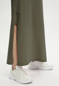 Langes olivgrünes Kleid mit einem Seitenschlitz, kombiniert mit weißen Sportschuhen, die drei blaue Streifen und eine strukturierte Sohle haben.