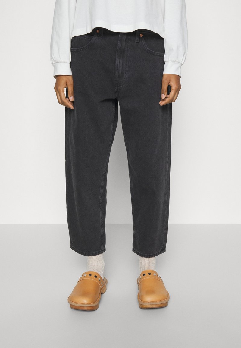 Pantalones de mezclilla negros de corte relajado, con cinco bolsillos y una longitud recortada, combinados con zuecos marrones y calcetines de color claro.