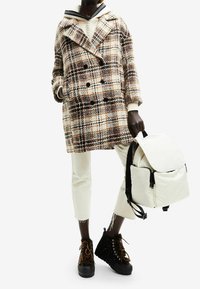 Cappotto di lana a scacchi nei toni crema, marrone e arancione; abbinato a pantaloni crema, stivaletti neri con rifiniture in pelliccia e uno zaino bianco.