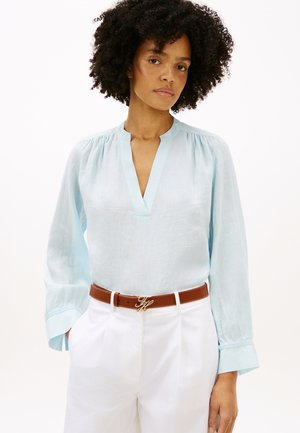 Γυναίκα με κατσαρά μαλλιά φοράει ανοιχτό μπλε μπλούζα με μακριά μανίκια και V-λαιμόκοψη, λευκό παντελόνι και καφέ ζώνη με χρυσή αγκράφα, στέκεται μπροστά σε λευκό φόντο.