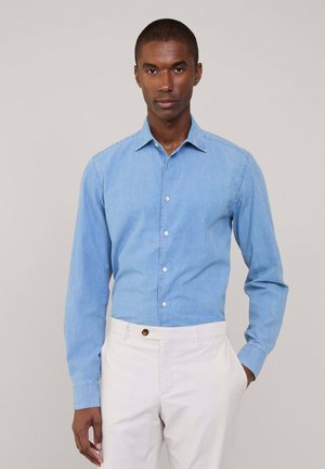 Camisa de mezclilla azul claro con cuello, mangas largas y cierre de botones. Combinada con pantalones blancos que tienen un acento de botón marrón.