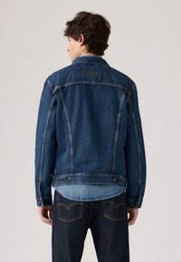 Donkerblauwe denimjas met een licht vervaagde afwerking, voorzien van een studsontwerp op de achterkant, knoopmanschetten en twee achterzakken.