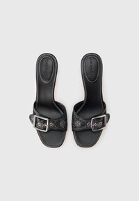 Fekete bőr papucs texturált pánttal és fém csattal. A talpon fehér varrással és sima talpfelülettel rendelkezik.