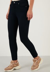 Jeans ajustados azul oscuro con una textura suave, cinco bolsillos y cierre de botón frontal, combinados con zapatillas marrón claro.