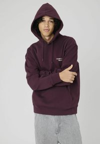 Sweat à capuche bordeaux avec poche avant et capuche à cordon réglable, portant un texte blanc indiquant "Create Your World". Porté avec un pantalon gris.