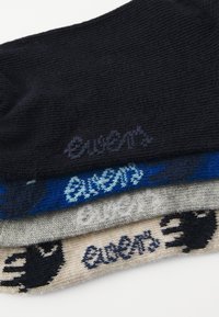 Cuatro pares de calcetines: negro, azul, gris y beige. Cada uno presenta un logo bordado diferente en el puño. Material de punto texturizado.