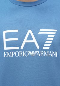 Μπλε βαμβακερό t-shirt με έντονο λευκό κείμενο "EA7" και λογότυπο "EMPORIO ARMANI". Στρογγυλή λαιμόκοψη με ομαλή υφή.