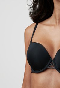 Svart vadderad bh med tunna justerbara band, scallopade kanter och en meshdesign i mitten för stöd. Slät, elastisk material.