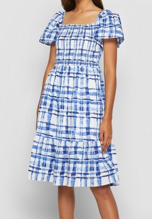 Robe à carreaux bleue et blanche avec des manches bouffantes, un décolleté carré et un bodice smocké, présentant un ourlet à volants en couches. Matériau en coton.