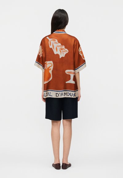 Γυναίκα στέκεται, φοράει υπερμεγέθη πουκάμισο χρώματος σκουριάς πορτοκαλί με λευκά αφηρημένα σχέδια και κείμενο στο κάτω μέρος, συνδυασμένο με μαύρα σορτς μέχρι το γόνατο και καφέ παπούτσια.