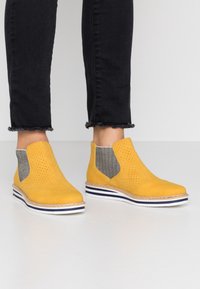 Żółte zamszowe botki za kostkę z perforowanym wzorem, szare tekstylne panele boczne i biała podeszwa z granatowymi i beżowymi pasami.