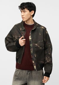 Νεαρός άνδρας που ρυθμίζει ένα φθαρμένο αδιάβροχο τζάκετ καμουφλάζ πάνω από ένα μπορντό πλεκτό πουλόβερ και ξεθωριασμένο γκρι τζιν, κοιτάζοντας προς το πλάι.