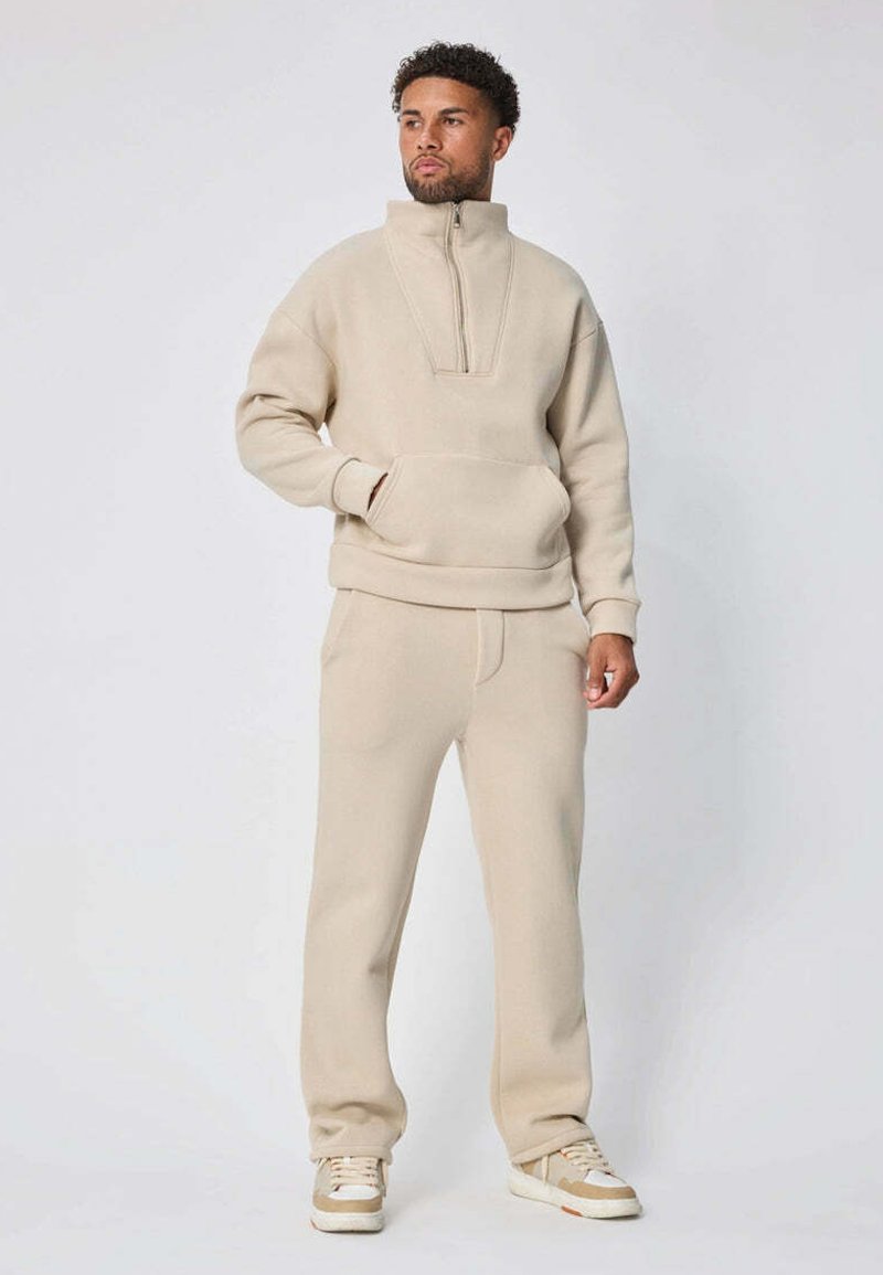 Homme debout portant un pull polaire beige avec une demi-fermeture éclair, un pantalon assorti et des baskets beiges, regardant légèrement sur le côté.