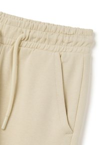 Pantalones de chándal beige con banda elástica en la cintura y cordón, bolsillos laterales y una textura suave. Diseño sencillo sin patrones visibles.