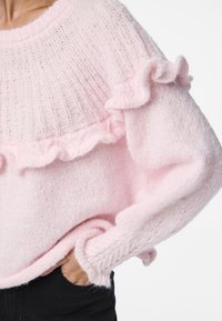 Pull en tricot rose clair avec une section supérieure côtelée et des volants le long de la partie médiane et des manches. Texture douce.