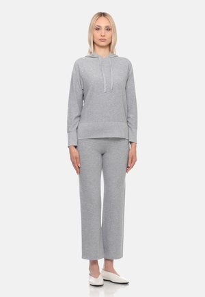 Donna bionda in piedi che indossa una felpa con cappuccio grigio chiaro e pantaloni larghi abbinati con scarpe basse bianche su uno sfondo semplice.