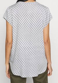 Osoba stojící s viditelným zadním pohledem, oblečená v bílém tričku s krátkým rukávem a černým geometrickým vzorem a olivově zelených kalhotách.