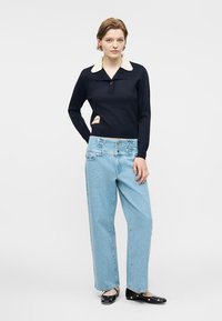 Jeune femme portant un haut noir à manches longues avec un col blanc, un jean taille haute à jambes larges bleu clair et des ballerines noires ornées de détails en forme de cœur.