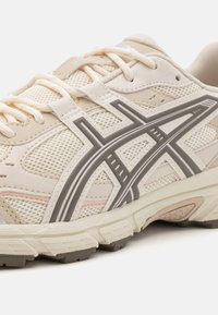 Sapata desportiva de cor bege clara com parte superior em malha, sola em borracha e detalhes cinzentos. Apresenta um design com atacadores e áreas texturizadas para aderência e suporte.