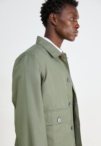 Olivgrön jacka med krage, knappstängning och framfickor med kontrasterande knappar; slät, slitstark tyg med en strukturerad passform.