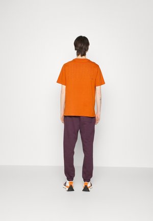 Orange bomullströja med korta ärmar och en baksidedesign med små perforeringar. Bärs med lila joggers och sneakers.