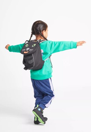Kind mit geflochtenem Haar, das einen türkisen Pullover, eine marineblaue Jogginghose und einen schwarzen, katzenförmigen Rucksack trägt, steht mit ausgebreiteten Armen.