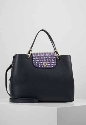 Sac à main en cuir noir avec poignées courtes, bandoulière détachable et rabat à motif avec un emblème doré circulaire.