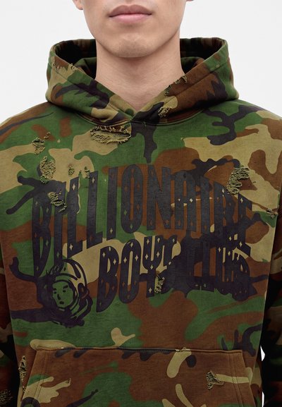 Young person wearing green and brown camouflage hoodie with distressed details and "Billionaire Boys Club" text and astronaut logo on front.