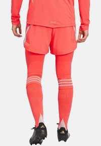 Heldere neonroze sportshorts en leggings van gestructureerde stof, met drie horizontale strepen bij de kuiten en zwarte voetbalschoenen.