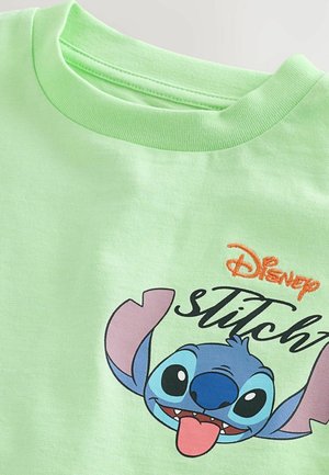 Gaiši zaļš kokvilnas t-krekls, kas rotāts ar grafiku ar multfilmu varoņa seju, kurai ir rozā ausis, zilas sejas iezīmes un melnas acis, ar uzrakstu "Disney Stitch".