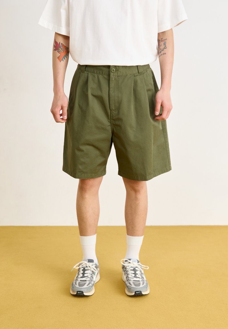 Groene katoenen shorts met een relaxed fit, voorzien van een knoopsluiting, zijzakken en een gladde textuur, gecombineerd met grijze sneakers en witte sokken.