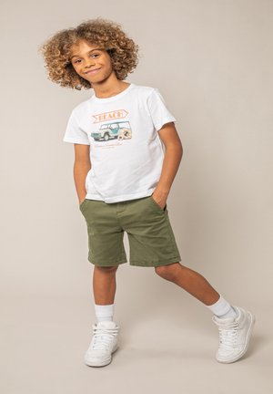 Enfant aux cheveux bouclés souriant, portant des baskets blanches, un t-shirt graphique blanc et un short vert olive, debout sur un fond beige les mains dans les poches.