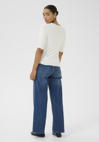 Hvid ribbet top med korte ærmer, bølgede kanter, parret med løst fit mørkeblå denim jeans, vist fra bagsiden.