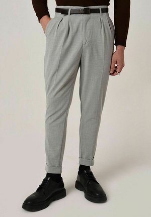 Pantalon gris plissé avec une coupe ajustée, doté d'une ceinture et d'ourlets retroussés, associé à des chaussures noires à lacets. Texture de tissu lisse.