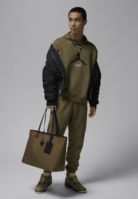 Felpa verde oliva con logo Jordan nero, pantaloni jogger coordinati, giacca bomber nera e una borsa a spalla marrone con motivi e accenti neri.