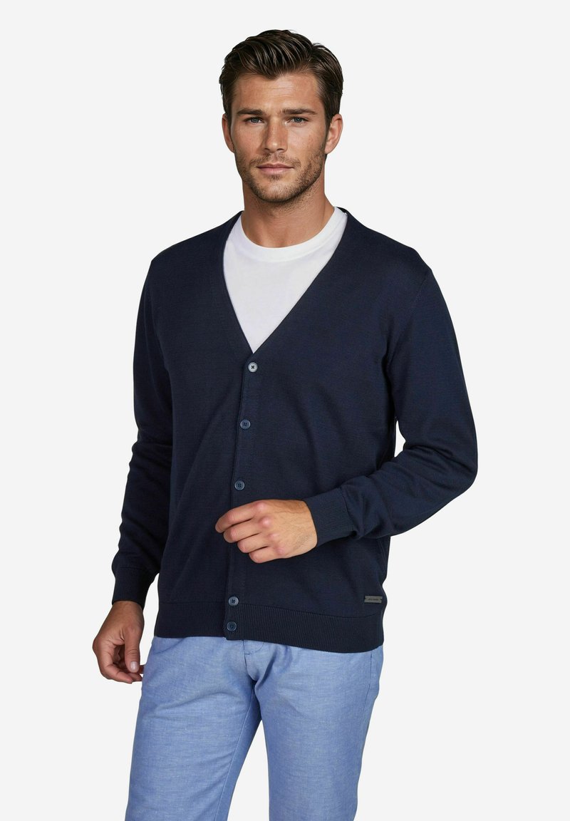 Marineblaue Strickjacke mit V-Ausschnitt, Vorderknöpfen und gerippten Bündchen sowie Saum. Kombiniert mit hellblauen Hosen.