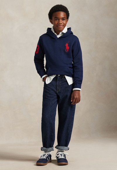 Junge trägt einen marineblauen Hoodie mit rotem Logo, darunter ein weißes Hemd, dunkle hochgekrempelte Jeans und marineblaue Sneakers mit weißen Schnürsenkeln, steht mit einer Hand in der Tasche.