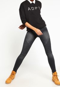 Sudadera negra con texto blanco "ADAPT", combinada con jeans skinny gris oscuro que presentan paneles de textura acanalada, y botas de tobillo marrón.