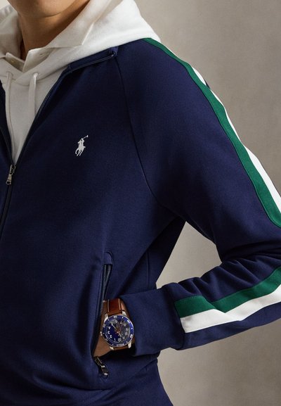 Homme portant une veste zippée bleu marine avec des bandes vertes et blanches sur les manches et un sweat à capuche blanc en dessous, montrant une montre-bracelet avec un bracelet en cuir marron.