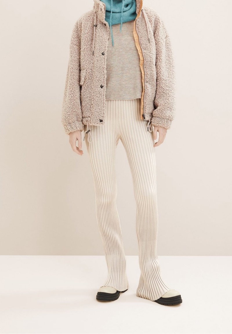 Personne portant une veste sherpa beige, un pull gris clair, un pantalon crème côtelé et des chaussures bicolores crème et noir, debout devant un fond neutre.