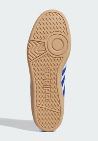 Suela exterior de goma con un diseño texturizado; cuenta con un elemento de diseño circular y la marca "adidas" en marrón a lo largo del lateral, con acentos en azul visibles.