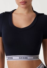 Top corto azul marino con mangas cortas, escote en forma de barco y banda elástica con la marca "GUESS" en blanco en el dobladillo. Tejido suave y elástico.