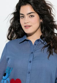 Camicia blu con bottoni, caratterizzata da un design ricamato con una corona e un cuore. La camicia ha un colletto e un tessuto liscio e leggero.